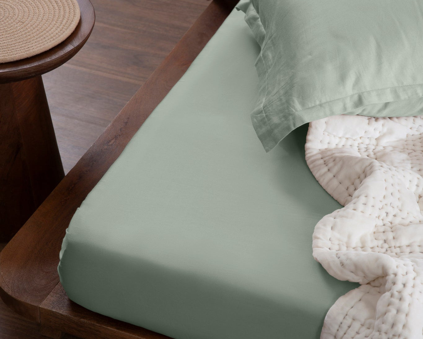 Aplito sage-green sheet and matching pillow cover on a wooden bed with an off-white quilt.