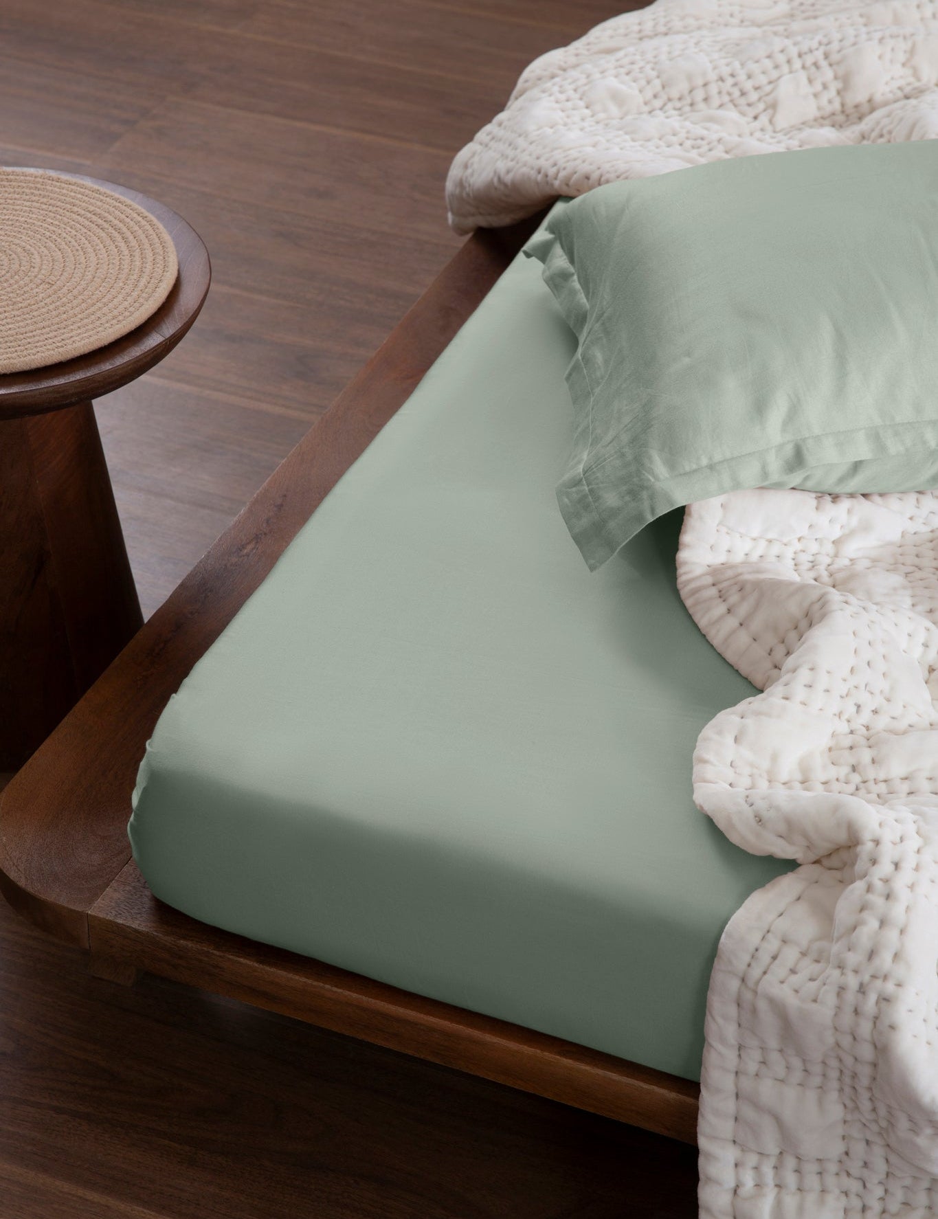 Aplito sage-green sheet and matching pillow cover on a wooden bed with an off-white quilt.