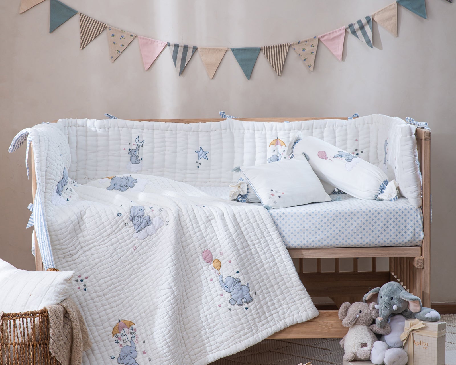 Aplito Little Tuskers Blue bedding with elephant motifs, a quilt and matching pillows displayed on a wooden cot.